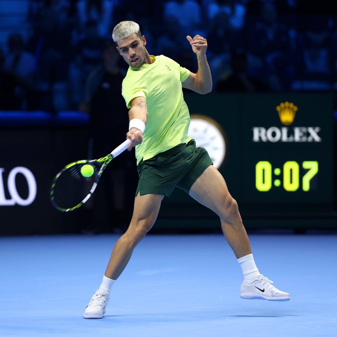 Carlos Alcaraz with platinum blonde hair wearing a lime green shirt and dark green shorts holds a tennis racket and hits a forehand shot toward the net on an indoor blue hard court during a match with a Rolex sponsor board and score display showing 0:07 in the background surrounded by spectators