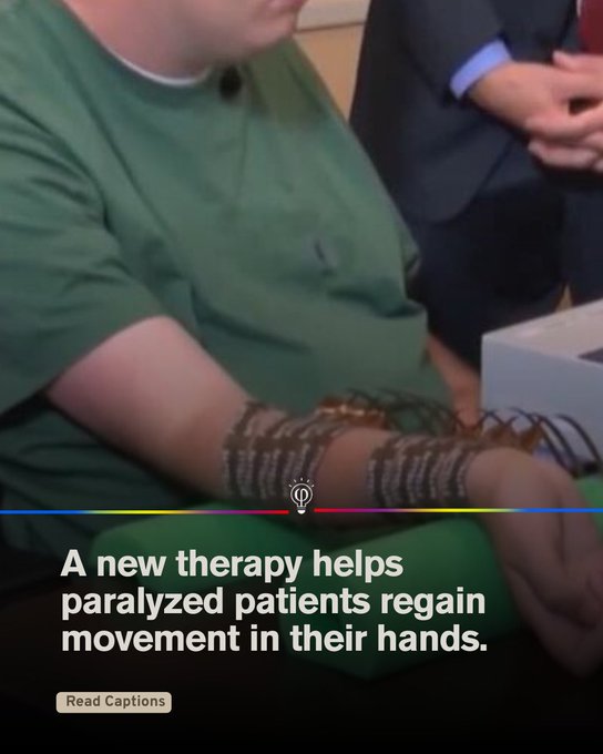A man wearing a green shirt sits with his right arm extended on a table, connected to multiple black wires and bracelets around his wrist and forearm, linked to a white device box. Another person in a blue suit stands nearby observing. The arm shows medical attachments for therapy. Blue and yellow colored bars with a circular icon appear at the bottom.