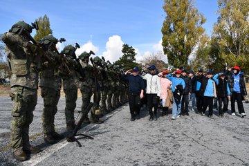 First image shows a line of soldiers in camouflage uniforms and helmets standing at attention with rifles on a paved area under a clear sky with trees and clouds in background, facing a group of civilians including adults and children in casual clothing walking towards them. Second image depicts two young girls wearing red caps and casual clothes inside a fire truck cab, one holding a radio and the other steering the wheel, with dashboard and equipment visible. Third image features a large group of soldiers in uniforms and civilians including children in blue clothing gathered in front of a banner with Turkish flags and a portrait of Ataturk, on an open ground with trees and vehicles nearby. Fourth image captures a group of people including military personnel in uniforms, suited individuals, and youth in red shirts posing together beside a green military helicopter with rotors, against a mountainous landscape.