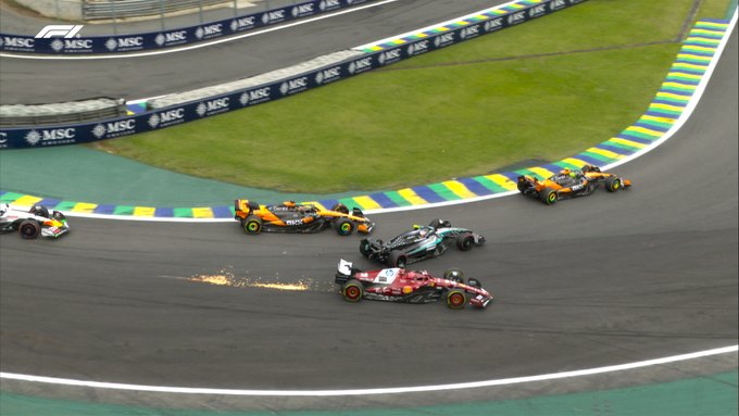 Aerial view of Formula 1 cars on a racetrack during a restart in the Brazil Grand Prix, with multiple vehicles including those driven by Oscar Piastri, Kimi Antonelli, and Charles Leclerc positioned three-wide approaching Turn 1, showing a collision where one car tangles and sustains visible damage, surrounded by colorful barriers, green grass, and sponsor banners.
