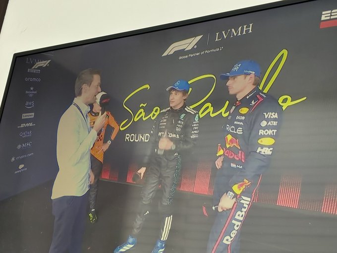 Large screen displays Formula 1 logo and LVMH sponsorship above text Sao Paulo Round. Three men stand below: left man in light shirt and pants with orange shoes speaks into microphone wearing lanyard. Center man in blue cap and suit with sponsor patches like Visa and Mercedes. Right man in blue cap and dark suit with patches like Red Bull and Rock. Background includes event branding and lighting.