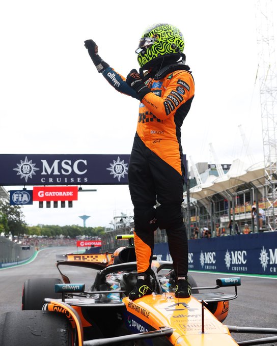 Lando Norris stands on a Formula 1 podium wearing an orange and black McLaren racing suit and a green patterned helmet, raising one arm in celebration. Behind him is a McLaren race car on the track with orange accents and sponsor logos. The background features a grandstand with MSC and Gatorade banners, FIA signage, and a covered spectator area under a partly cloudy sky.