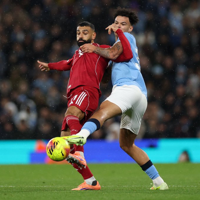 Two soccer players engage in a tackle on a green field during a match in a crowded stadium. The player in a red kit with white stripes reaches with his right arm while extending his left leg, wearing red and white cleats. The opposing player in a light blue kit with dark blue socks leans forward with tattoos visible on his arm, wearing white shorts and blue cleats. A yellow soccer ball is positioned between their legs near the touchline. Spectators fill the stands in the background under stadium lights.