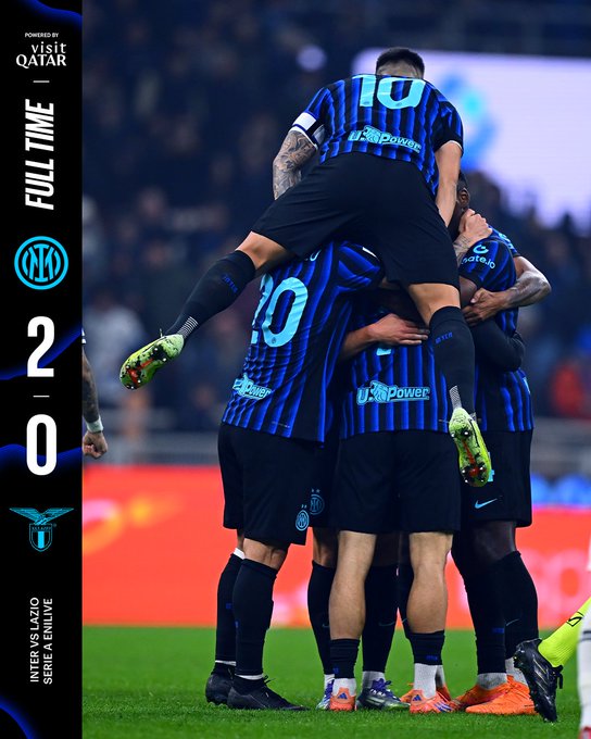 Several male soccer players in blue and black striped jerseys and shorts, with numbers like 10 visible, are stacked in a celebratory pile on a green grass field under stadium lights. The player at the bottom kneels with arms extended, while others jump on his back, arms raised in triumph. A vertical banner overlay shows FULL TIME INTER 2 LAZIO 0 with team logos including the Inter snake emblem and Lazio eagle, plus sponsor Visit Qatar and Qatar Airways logos. The background includes a blurred stadium crowd and field markings.