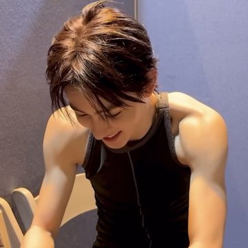 First image: Young man with short brown hair wearing black tank top smiling while seated leaning forward in room with blue walls and white chair. Second image: Same man holding iPhone 13 in hand wearing green tank top seated in similar room setup. Third image: Man outdoors on grassy area with rocks wearing white tank top white hooded towel with C logo around neck sunglasses hanging from towel gray pants sitting relaxed.