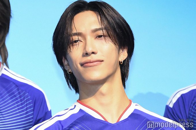 A young man with long dark hair parted in the middle and wearing small hoop earrings smiles slightly while dressed in a blue soccer jersey with red and white accents. He stands among other individuals in similar blue uniforms against a bright blue background with a mountain graphic. The photo is credited to Modelpress.
