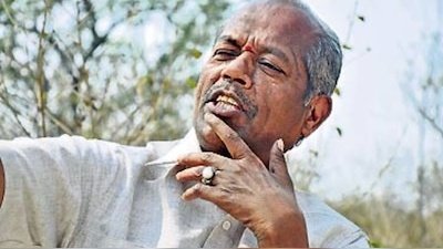 An elderly man with short gray hair and a mustache sits outdoors under a clear sky with green trees in the background. He wears a light-colored traditional shirt and has a thoughtful expression with his hand touching his chin. A large ring is visible on his finger.