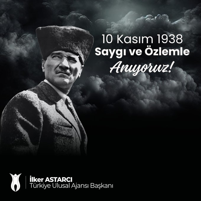 Black-and-white portrait of Mustafa Kemal Atatürk wearing a formal suit, overcoat, and traditional hat, facing forward with a serious expression. Dark cloudy background evokes solemnity. Prominent white text overlay states 10 Kasım 1938 at the top and saygı ve özlemle Anıyoruz below. Bottom features logos of Türkiye Ulusal Ajansı with a tulip emblem and İker Atarcanı Başkanı in white text.