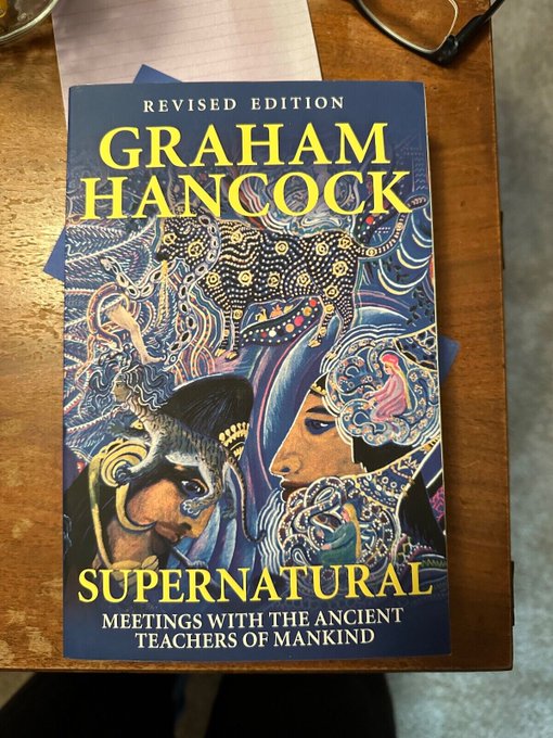 Book cover titled Supernatural Meetings with the Ancient Teachers of Mankind by Graham Hancock in revised edition, featuring intricate blue and yellow artwork of masked ancient figures with elaborate headdresses and patterns on a wooden table surface alongside glasses and papers.