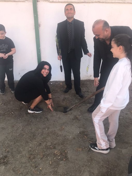 A group of adults and children stand near a white wall with green poles, one man in a suit holds an umbrella, a woman in a black hoodie and pants squats holding a shovel next to a girl in white outfit and pants who holds a shovel sticking into a dirt hole, another man in suit stands nearby, children in casual clothes observe. Several people including young adults and children in casual attire like hoodies and sneakers gather around a small planted sapling in dirt, one person pours water from a bottle onto the sapling, others hold shovels or stand watching. A small evergreen sapling planted in paved area near parked white and orange cars, a paper sign attached to it reads DİKİŞ ALİ ÇETİN KIZLARIN GÜCÜ in Turkish.