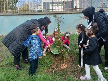 First image shows several young children wearing colorful party hats and casual clothes, assisted by adults in headscarves and jackets, kneeling around a freshly planted sapling in a bucket-filled hole on grassy ground near a blue wall and fence. Second image depicts a man and woman helping two young boys in dark clothing pour soil from a green bucket into a hole beside a small plant, with more grass and a fence in the background. Third image captures a large group of young children in various outfits including dresses and jackets, standing with several adults in headscarves and casual wear, in front of a brown backdrop banner featuring clock faces and handprints. Fourth image features a woman in formal attire standing with eight young children in school uniforms and casual clothes, positioned before a large brown banner displaying 1937-1938 dates, infinity symbols made of hands, and clock motifs against a building wall.