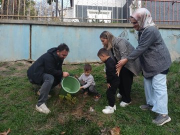 First image shows several young children wearing colorful party hats and casual clothes, assisted by adults in headscarves and jackets, kneeling around a freshly planted sapling in a bucket-filled hole on grassy ground near a blue wall and fence. Second image depicts a man and woman helping two young boys in dark clothing pour soil from a green bucket into a hole beside a small plant, with more grass and a fence in the background. Third image captures a large group of young children in various outfits including dresses and jackets, standing with several adults in headscarves and casual wear, in front of a brown backdrop banner featuring clock faces and handprints. Fourth image features a woman in formal attire standing with eight young children in school uniforms and casual clothes, positioned before a large brown banner displaying 1937-1938 dates, infinity symbols made of hands, and clock motifs against a building wall.