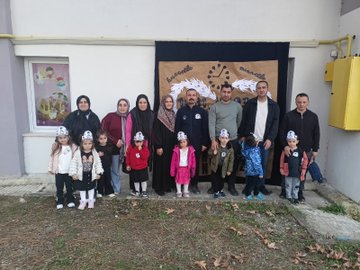 First image shows several young children wearing colorful party hats and casual clothes, assisted by adults in headscarves and jackets, kneeling around a freshly planted sapling in a bucket-filled hole on grassy ground near a blue wall and fence. Second image depicts a man and woman helping two young boys in dark clothing pour soil from a green bucket into a hole beside a small plant, with more grass and a fence in the background. Third image captures a large group of young children in various outfits including dresses and jackets, standing with several adults in headscarves and casual wear, in front of a brown backdrop banner featuring clock faces and handprints. Fourth image features a woman in formal attire standing with eight young children in school uniforms and casual clothes, positioned before a large brown banner displaying 1937-1938 dates, infinity symbols made of hands, and clock motifs against a building wall.
