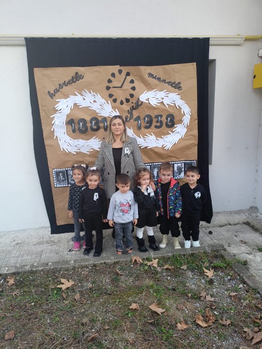 First image shows several young children wearing colorful party hats and casual clothes, assisted by adults in headscarves and jackets, kneeling around a freshly planted sapling in a bucket-filled hole on grassy ground near a blue wall and fence. Second image depicts a man and woman helping two young boys in dark clothing pour soil from a green bucket into a hole beside a small plant, with more grass and a fence in the background. Third image captures a large group of young children in various outfits including dresses and jackets, standing with several adults in headscarves and casual wear, in front of a brown backdrop banner featuring clock faces and handprints. Fourth image features a woman in formal attire standing with eight young children in school uniforms and casual clothes, positioned before a large brown banner displaying 1937-1938 dates, infinity symbols made of hands, and clock motifs against a building wall.