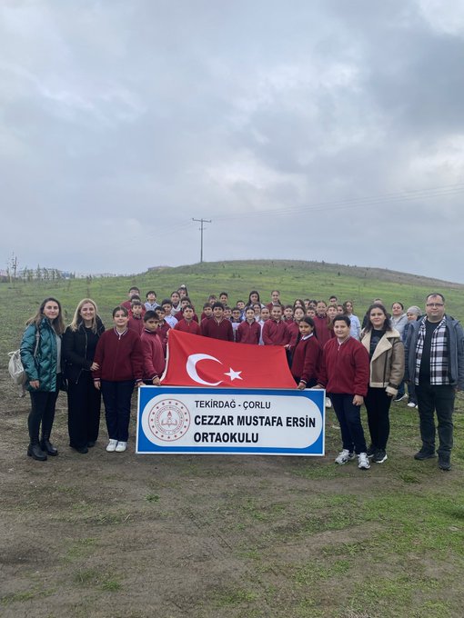 Group of students and adults in school uniforms and casual clothes stand on grassy hill holding Turkish flag and blue banner with school name Cezzar Mustafa Ersin Ortaokulu, cloudy sky and fence in background. Collage shows children in red and white uniforms planting small green sapling in soil with yellow plaque featuring trees and hands holding plant labeled Cezzar Mustafa Ersin Ortaokulu Ali, multiple angles of activity. Young tree sapling with branches in soil next to yellow plaque labeled Başöğretmen Mustafa Kemal Atatürk with tree icons and hands holding plant, children and woman in background wearing casual clothes.