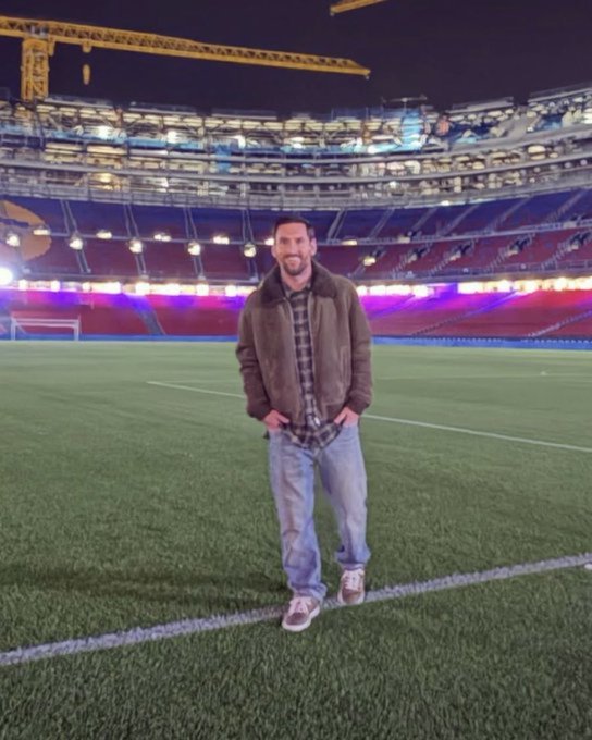 Lionel Messi stands on the green field near the center line, dressed in a brown shearling jacket over a plaid shirt, blue jeans, and white sneakers. The Spotify Camp Nou stadium surrounds him with empty tiered seats illuminated by vibrant purple and red lights. Construction cranes rise in the background against the night sky, and the goalpost is visible in the distance.