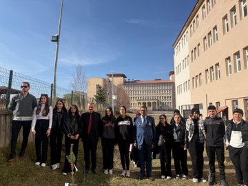 First image shows a man in a black suit and red tie kneeling to plant a small pine sapling with a shovel near a wire fence under a clear blue sky with the sun visible. Second image depicts a group of about 15-20 adults and young people including men in suits and casual wear and women in various outfits standing together on grass in front of a beige school building holding red flowers. Third image features six young children in orange uniforms with white shirts kneeling in a circle around a newly planted sapling each holding a white paper while smiling at the camera on grassy ground. Fourth image is a collage of four scenes showing groups of adults and children in formal and casual clothes digging and planting trees with shovels near school buildings and fences under sunny skies.
