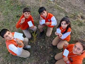 First image shows a man in a black suit and red tie kneeling to plant a small pine sapling with a shovel near a wire fence under a clear blue sky with the sun visible. Second image depicts a group of about 15-20 adults and young people including men in suits and casual wear and women in various outfits standing together on grass in front of a beige school building holding red flowers. Third image features six young children in orange uniforms with white shirts kneeling in a circle around a newly planted sapling each holding a white paper while smiling at the camera on grassy ground. Fourth image is a collage of four scenes showing groups of adults and children in formal and casual clothes digging and planting trees with shovels near school buildings and fences under sunny skies.