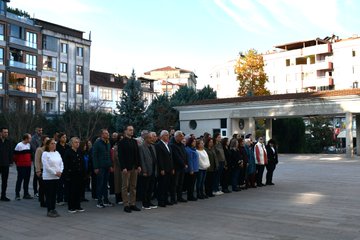 First image shows a bronze statue of a man in military uniform standing at attention with a Turkish flag nearby, in front of a modern beige building with large glass windows and a municipal emblem, a group of about 20 adults in casual and formal attire standing in a line facing the statue on a paved courtyard under clear skies. Second image depicts a similar group of around 25 adults, including men and women in coats and jackets, lined up in front of the same building with autumn trees in yellow and orange foliage visible to the side, under sunny conditions. Third image features the statue and flag again, with a smaller group of about 15 people, some in headscarves, standing respectfully in front of the glass-fronted entrance and courtyard. Fourth image shows a larger gathering of over 30 adults in various outfits including jackets and dresses, positioned in rows on the pavement in front of the multi-story town hall building with evergreen trees and blue sky in the background.