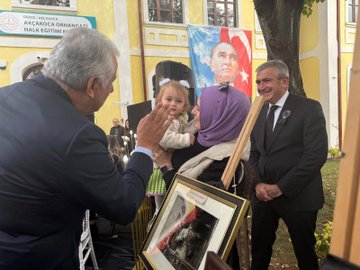 First image shows group of people including men in suits and women in headscarves standing near large Turkish flag with crescent and star against yellow building with trees in background. Second image captures elderly man in suit holding baby girl with headband while another man in suit smiles and touches her cheek near framed portrait of Mustafa Kemal Atatürk and Turkish flag. Third image features men in military and civilian attire standing before row of black-draped frames containing portraits on grassy area with yellow flowers. Fourth image displays officials near speaker setup and easels with portraits including large banner of Mustafa Kemal Atatürk face against yellow building.