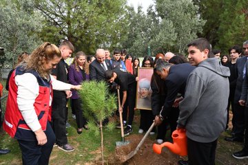 Group of people including men in suits, women in professional attire, young men and women in casual clothes, and officials standing around a freshly dug hole in grassy area surrounded by olive and pine trees, holding shovels and a portrait frame of a man, with a small pine sapling in a pot nearby. Another scene shows participants pouring water from a watering can onto the planted sapling using a red can, with shovels inserted in the soil beside the tree. Third image depicts individuals in similar attire planting another sapling, one person touching the tree branch, holding the portrait, and using a shovel near the hole. Fourth image shows diverse group of men and women, some wearing headscarves, seated around an oval table in a room with large windows, plates of food and drinks on the table, engaged in discussion.