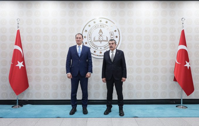 First image shows two men in dark suits standing side by side in front of a light gray wall with the emblem of the Turkish Ministry of National Education centered, flanked by two Turkish flags on stands. The man on the left has short dark hair and wears a blue tie, while the man on the right has glasses and a gray tie. The floor is blue carpet with a modern interior setting. Second image depicts two men in suits shaking hands over a table with water bottles and documents, holding red-bound agreements labeled Turkiye Malta in white text on screens behind them showing Turkish and Maltese flags. The setting is a formal conference room. Third image features a group of six people seated in an ornate room with sofas and a wooden table, including men in suits and a woman in a headscarf, with Turkish and another flag, a painting of children on the wall, and patterned carpet.