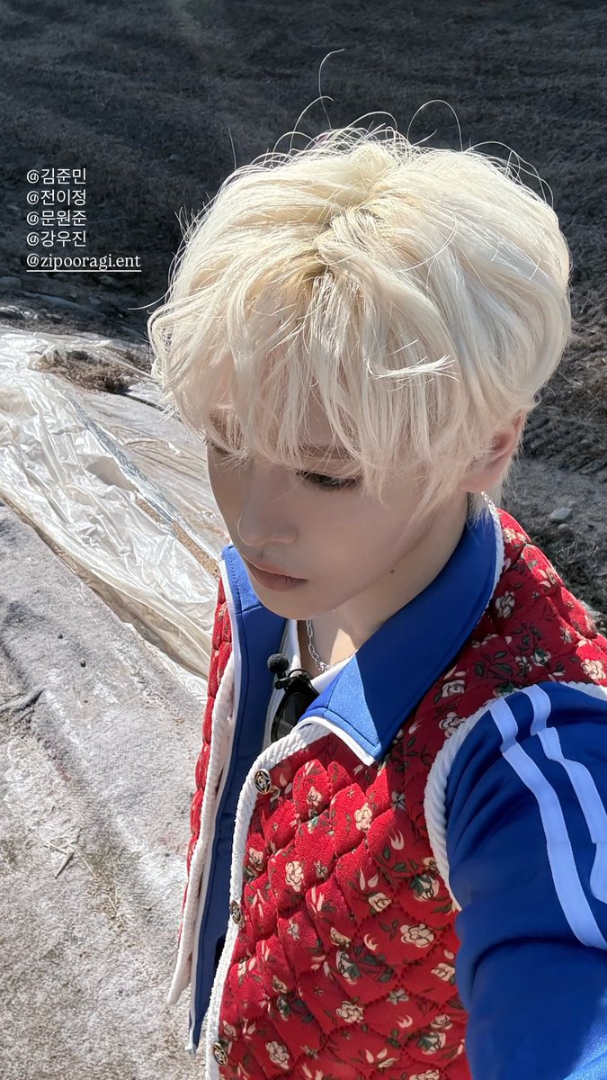 A young man with short blonde hair and fair skin poses sideways, looking to the side with a neutral expression. He wears a quilted jacket in red and multicolored floral patterns with blue sleeves and collar, featuring white stripes on the arms. The background is plain and neutral.