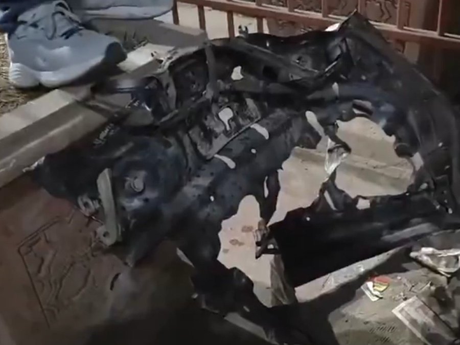 Charred remains of a car with extensive fire damage visible on the body and interior exposed through broken windows and doors next to a metal fence and ground debris including shoes and papers.