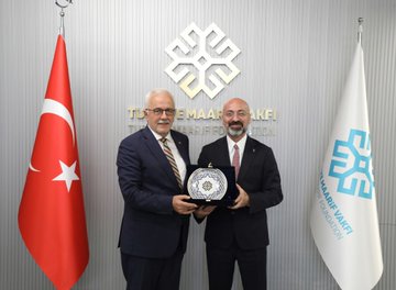 First image shows two men in suits sitting at a wooden table with documents, one older man with glasses signing papers, the other bald man also signing, backdrop with blue Türkiye Maarif Vakfı logo. Second image depicts the two men standing, each holding a red-bound book with gold emblem, wearing suits and ties, backdrop with white banner and blue logo. Third image features the two men standing with Turkish and white flags, holding a round plaque with geometric design, suits and ties, wall with Türkiye Maarif Vakfı logo. Fourth image shows a group of eight people including men and one woman in formal attire standing together, some in suits, woman in skirt and headscarf, backdrop with flags and logo.