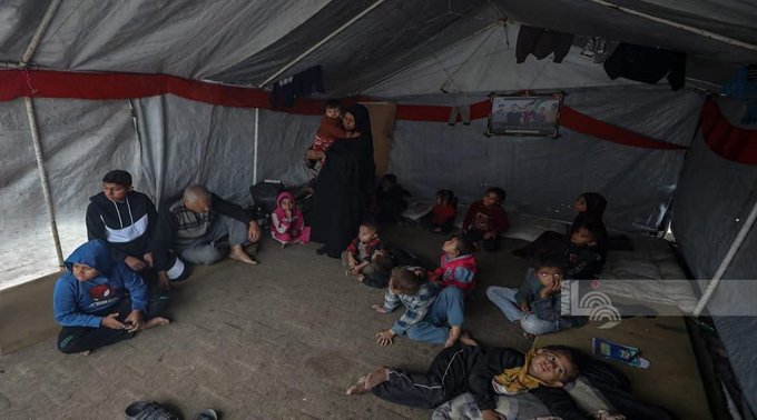 First image shows a group of children and adults sitting inside a large tent with white and red fabric walls and a hanging cloth. Several young children in colorful clothes like pink jackets and blue outfits sit on the floor holding hands or eating, surrounded by older women in black abayas. A boy in a blue jacket and another in a striped shirt are visible among the group gathered closely together on mats.