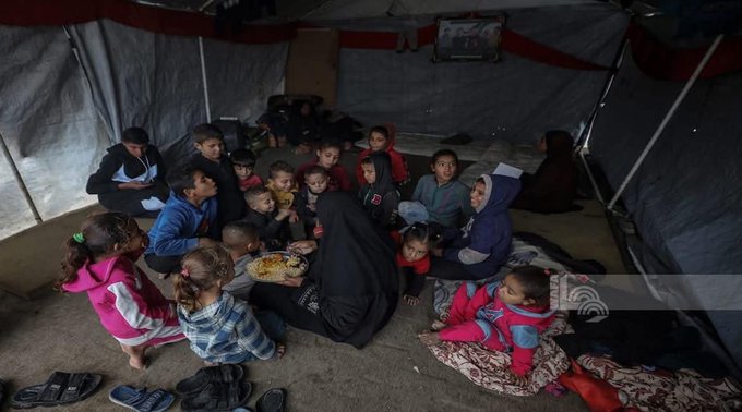 First image shows a group of children and adults sitting inside a large tent with white and red fabric walls and a hanging cloth. Several young children in colorful clothes like pink jackets and blue outfits sit on the floor holding hands or eating, surrounded by older women in black abayas. A boy in a blue jacket and another in a striped shirt are visible among the group gathered closely together on mats.