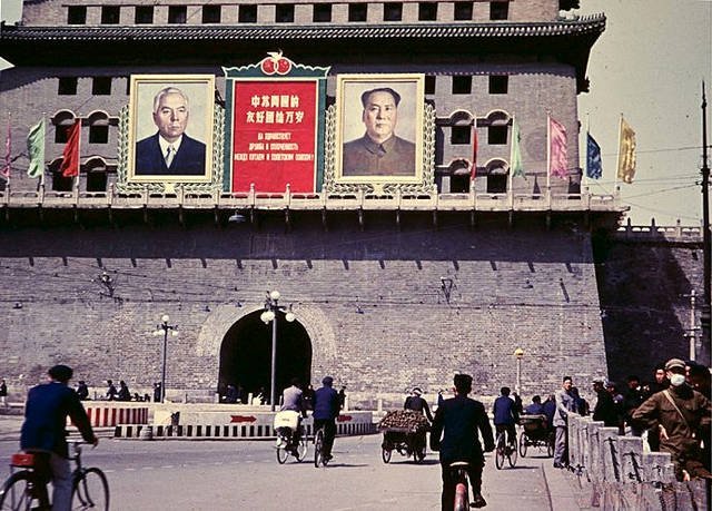 北京天安门的彩色历史照片，悬挂大型横幅，展示毛泽东及另一位领导人的肖像，标有中文文字。红旗和绿色装饰悬挂在建筑上。一群身着1950年代服装的人群，包括穿西装的男人和穿裙子的女人，在前景靠近栏杆和路灯旁行走和骑自行车。