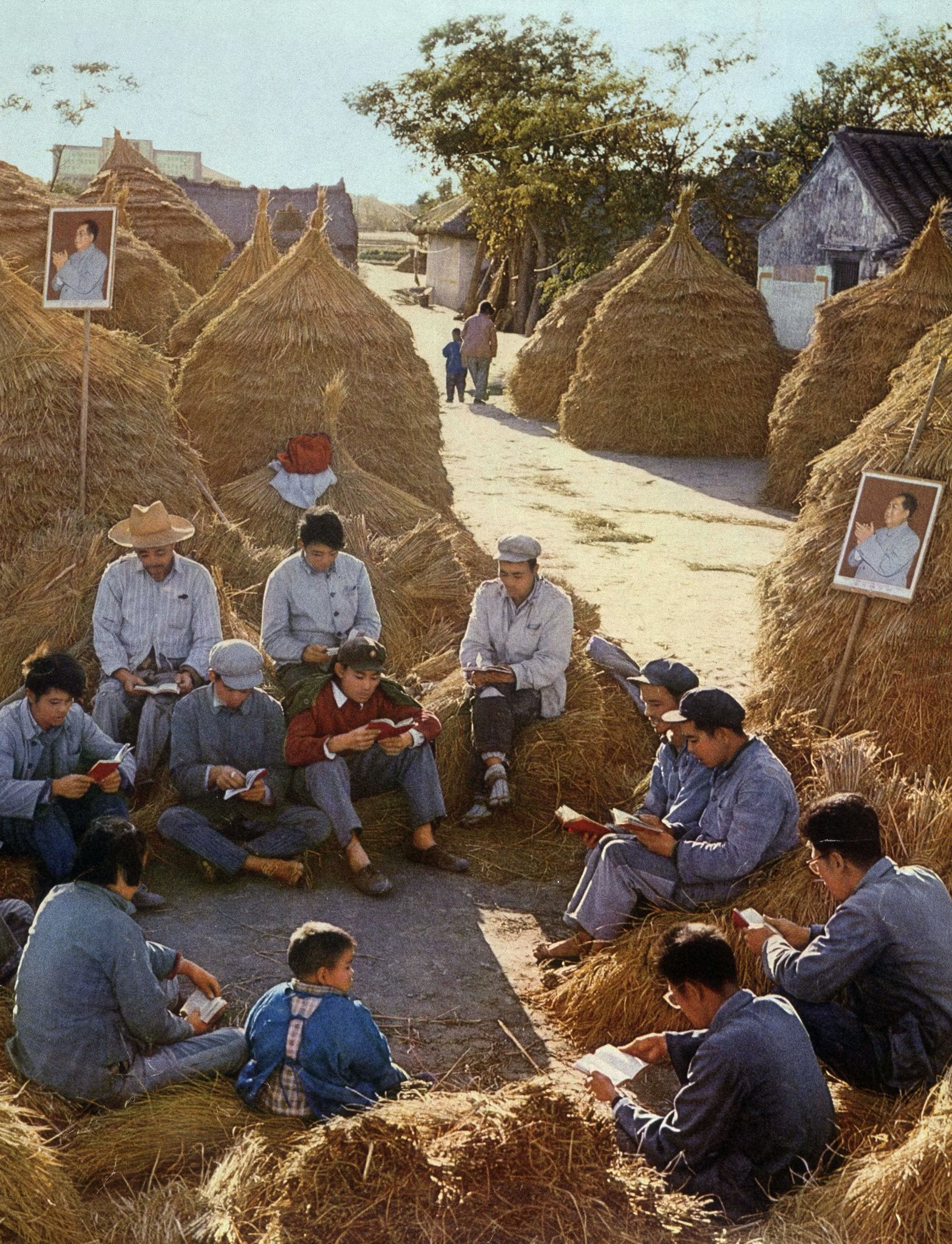 Group of people in traditional rural Chinese village setting with thatched haystack houses along a dirt path, several men and women in simple 1960s-era clothing including hats and jackets sitting on ground surrounded by hay bales, reading books attentively, a young boy in blue jacket also reading, framed black-and-white portraits of individuals mounted on some haystack walls, trees and open sky in background.
