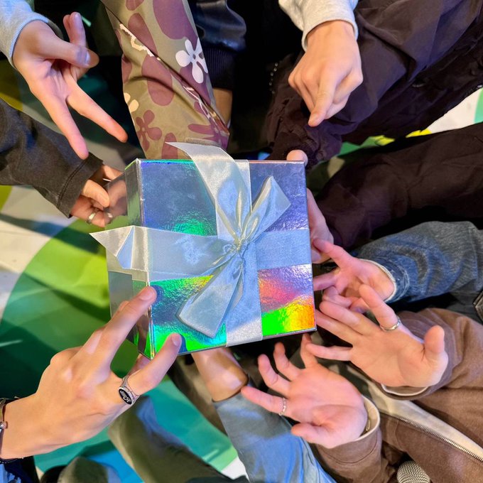 Multiple hands from people wearing casual clothing including patterned pants, sweaters, and rings extend towards a shiny silver gift box wrapped with a white ribbon, positioned on a green and colorful background.