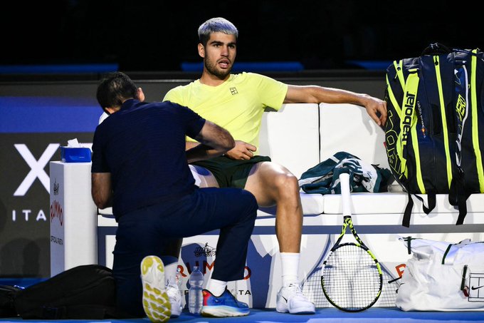 Carlos Alcaraz with silver-dyed hair sits on a white bench wearing a yellow shirt and green shorts, extending his arm while a man in a light blue shirt kneels beside him adjusting something on his leg. Tennis rackets, black and green bags, water bottles, and white bags are placed around the blue court floor with white lines and a sponsor logo visible in the background.