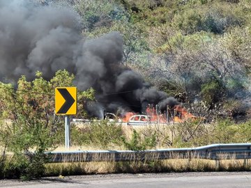 First image shows three individuals two in tactical gear with rifles and one central figure with blurred face wearing a gray Champion hoodie hands cuffed behind back standing against a plain wall. Second image depicts a rural road with thick black smoke rising from multiple burning orange vehicles near a chain link fence surrounded by green grass trees and a clear blue sky a person in dark clothing walks away carrying a bag. Third image features two green military style vehicles with armed personnel in camouflage one with blue lights on top parked on a dirt road beside desert landscape and a sign reading CONTROL. Fourth image illustrates a curved road with several burning cars emitting heavy black smoke yellow road sign indicating a curve vegetation and distant hills in background.
