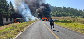 First image shows three individuals two in tactical gear with rifles and one central figure with blurred face wearing a gray Champion hoodie hands cuffed behind back standing against a plain wall. Second image depicts a rural road with thick black smoke rising from multiple burning orange vehicles near a chain link fence surrounded by green grass trees and a clear blue sky a person in dark clothing walks away carrying a bag. Third image features two green military style vehicles with armed personnel in camouflage one with blue lights on top parked on a dirt road beside desert landscape and a sign reading CONTROL. Fourth image illustrates a curved road with several burning cars emitting heavy black smoke yellow road sign indicating a curve vegetation and distant hills in background.