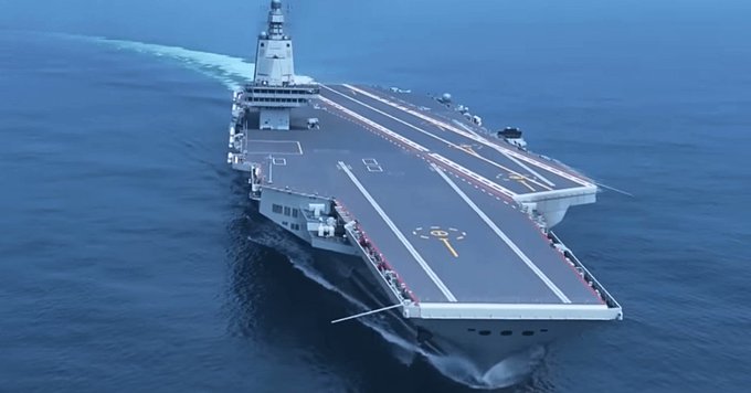 Aerial view of a large gray aircraft carrier sailing on the ocean with white waves trailing behind. The deck features angled runways marked with yellow lines and a circular landing area. Multiple aircraft are positioned on the deck including fighter jets with red stars. The ship has a prominent island superstructure on the starboard side with radar arrays and antennas. The hull displays the number 18 in white.