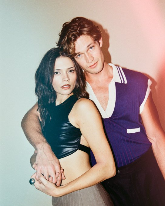 A young woman with long dark hair wearing a black leather crop top and matching skirt stands closely with a young man who has tousled brown hair and wears a blue sweater vest over a white shirt and black pants. They embrace with arms around each other and hands clasped holding a ring. The background is plain and the image has a warm filter.