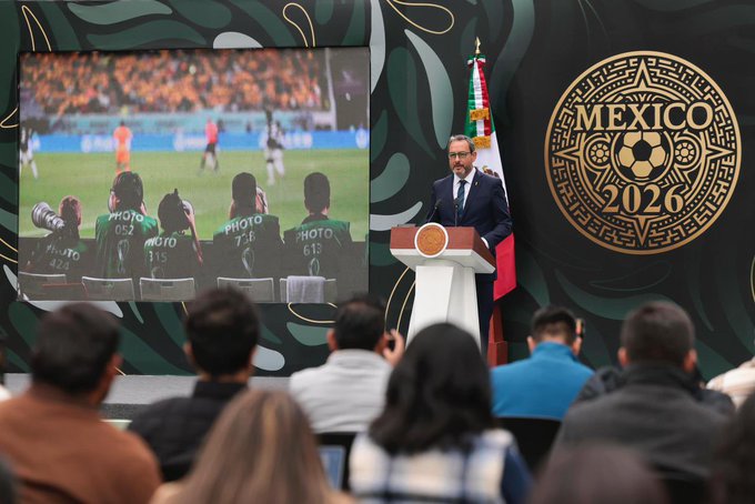 A split-view image shows a formal conference setting with a large screen displaying a soccer match on a green field with orange-uniformed players and a ball in play. In the foreground a man in a dark suit stands at a wooden podium with a microphone addressing an audience of seated and standing people including men and women in casual attire. The background features green decorative elements a Mexican flag and a large golden emblem with Mexico 2026 text encircled by intricate designs.