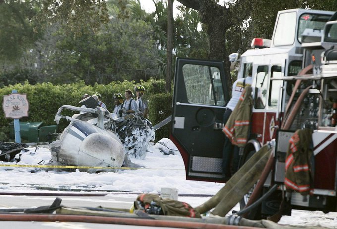 Wreckage of a small white airplane lies crashed on a grassy area near a road with white foam covering parts of it surrounded by yellow caution tape firefighters in uniforms stand near an open door of a red fire truck with hoses and equipment visible trees and bushes in the background a sign reading Inn on the Green is partially seen.