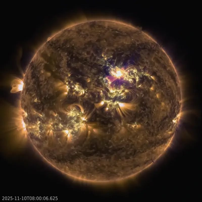 A spherical celestial body resembling the Sun with a glowing orange and yellow surface, featuring multiple dark sunspots clustered in one region, bright white plasma flares erupting from the sunspots, and purple and magenta coronal loops arching above the surface, captured in a high-contrast image highlighting solar magnetic activity.