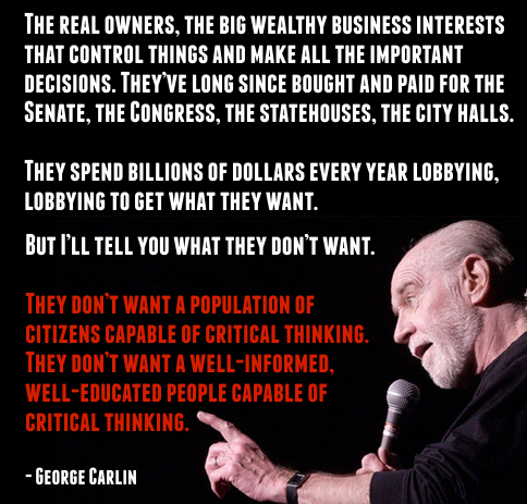 An elderly man with white hair and a beard stands on stage speaking into a handheld microphone. He wears a dark shirt and gestures with his right hand extended forward while pointing. The background is dimly lit with a blurred audience visible.