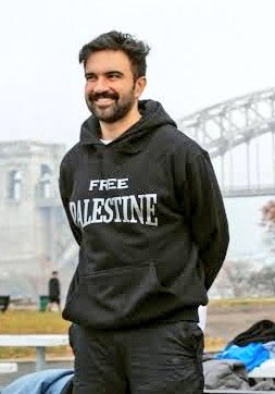 A man with short dark hair and a beard smiles while wearing a black hoodie with FREE PALESTINE printed in large white letters across the chest, standing in an outdoor park area with a large suspension bridge visible in the background under a foggy sky, other people and benches present nearby.