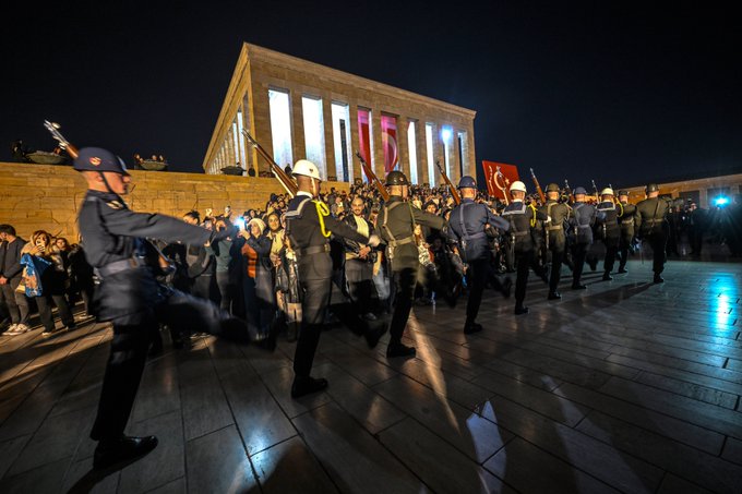 Group of uniformed military personnel in formal attire with rifles and yellow sashes stand in formation on a paved area at night in front of a large illuminated white stone mausoleum building with multiple columns and a Turkish flag displayed prominently on the facade surrounded by spectators.