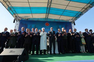 First image shows a man in suit and sunglasses standing at a podium with Turkish flag emblem, raising his right hand in gesture, against blue backdrop with text 11 Kasım Millî Ağaçlandırma Günü and green leaf motifs. Second image depicts several suited men including one identified as Recep Tayyip Erdoğan pouring water from green watering can onto young pine tree sapling, surrounded by applauding crowd with children in white outfits nearby. Third image features group of men in suits and traditional attire standing together on green stage under blue canopy with Turkish flag, some raising hands. Fourth image shows suited man using green shovel to plant tree sapling in soil, with other suited individuals and orange-clad workers around young pine trees and watering cans under sunny sky with stage in background.