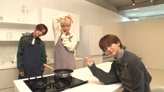 Three young men stand in a bright modern kitchen with white cabinets glass shelves holding dishes and potted plants on counters one with reddish hair wears a green apron over dark clothes and holds a wooden spoon stirring a black pot on an electric stove another with blonde hair in a white apron over light clothing raises both arms playfully the third with brown hair in a gray apron over dark shirt points forward with one hand they all smile engagingly the floor is light wood and walls are neutral tones
