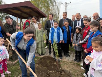 First image shows two red fire trucks with Turkish flags parked side by side in an open area with a green banner reading Sevdamız Toprak İmzamız Yeşil Vatan featuring children and adults wearing green hats standing in front smiling. Second image depicts a group of men in suits and casual clothes gathered around a young pine sapling being planted in soil with shovels and a blue banner in background under cloudy sky. Third image captures women and children in colorful clothing digging and planting small trees near a wooden pavilion with adults observing. Fourth image features adults and children in sportswear and vests kneeling and using shovels to plant a sapling in grassy soil area.