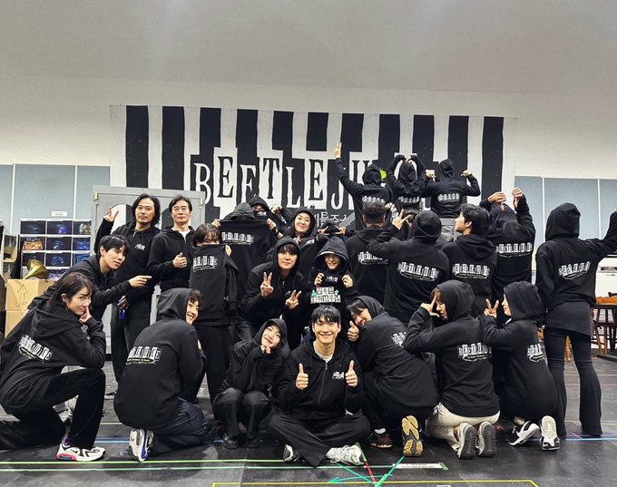 Group of approximately 20 young adults mostly Asian wearing black hoodies and pants with BEATLES text on chest and sleeves some with hoods up posing energetically in a semi-circle some making peace signs thumbs up or V signs casual smiles and excited expressions in front of large black and white vertical striped backdrop labeled BEATLES setting resembles indoor stage or gym with gray walls cardboard boxes and stage flooring visible