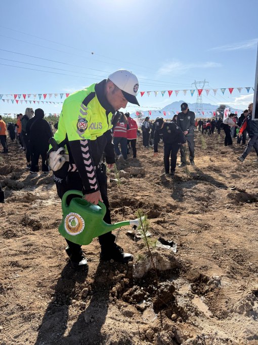 First image shows two young children in warm clothing and colorful boots kneeling on dry soil planting a small tree sapling together with focused expressions. Second image captures a woman in green hijab and yellow raincoat assisting a child in white cap and yellow raincoat planting a sapling using a watering can near stakes and other young trees on a hillside. Third image features a group including a man in blue cap two women in white tops and a girl in school uniform digging and planting trees on a sunny hillside with more participants in background. Fourth image depicts a police officer in uniform and white cap watering a newly planted sapling with a green watering can amid a crowd of people under Turkish flags on a clear day.